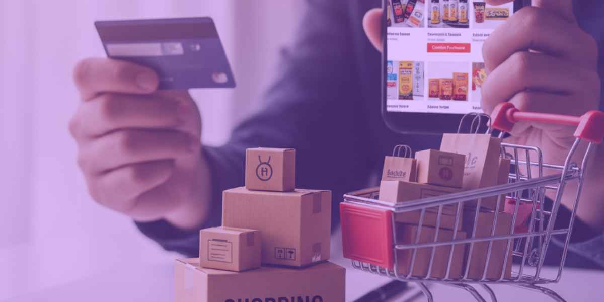 Uma pessoa mexendo no celular e, na outra mão, segurando um cartão de crédito. Ao lado, caixas de compras e um carrinho com compras dentro.