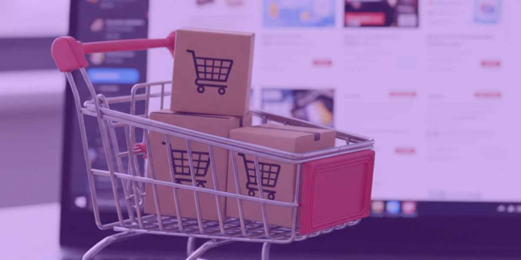 Um carrinho de compras com algumas caixas dentro e, ao fundo, um notebook exibindo uma plataforma de compras digital.