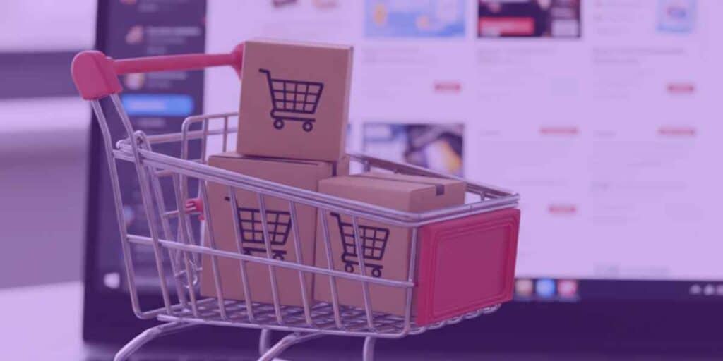 Um carrinho de compras com algumas caixas dentro e, ao fundo, um notebook exibindo uma plataforma de compras digital.